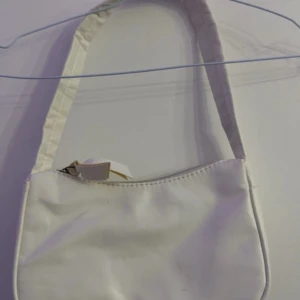 Vit axelväska i syntetmaterial - Säljer en vit axelväska med dragkedja. Väskan har en enkel design och är tillverkad i syntetmaterial. Perfekt för dig som vill ha en clean och minimalistisk look. Den är lite svarta streck men annars inget knappt använd
