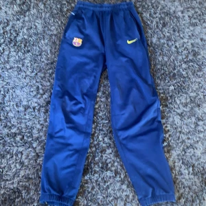 Blå Nike Dri-Fit Barcelona träningsbyxor - Säljer ett par blå träningsbyxor från Nike med Barcelona-logga och Dri-Fit material. Byxorna har resår i midjan och benslut samt gul Nike-logga på framsidan. Perfekta för fotbollsträning eller sportiga dagar.