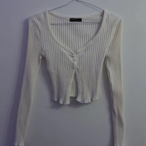 Vit ribbad croppad tröja från SHEIN - Säljer en vit ribbad croppad tröja från SHEIN med lång ärm och v-ringning. Tröjan har knappar framtill och en lätt volangkant nedtill. Perfekt att styla över ett linne eller t-shirt.