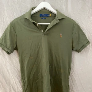 Olivgrön pikétröja från Polo Ralph Lauren - Säljer en klassisk olivgrön pikétröja från Polo Ralph Lauren med korta ärmar och liten broderad logga på bröstet. Tröjan har krage och knappar framtill. Perfekt för en stilren och avslappnad look.
