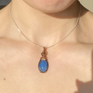 Handgjord berlock med blå sten och bronsfärgad tråd - Säljer ett elegant halsband med en oval blå sten infattad i bronsfärgad metalltråd. Hänget har fina detaljer och en unik tråddesign upptill. Perfekt för dig som vill ha något som sticker ut.