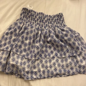 Vit och blå mönstrad kjol från H&M - Supersöt kortkjol från H&M i vitt med blått grafiskt mönster. Kjolen har smock i midjan och är luftig med volangdetalj nertill. Perfekt för varma dagar och enkel att matcha med en enkel topp.❤️