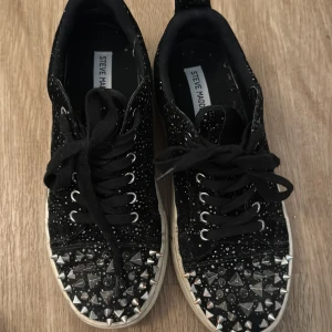 Svarta sneakers med nitar och glitter - Säljer ett par svarta sneakers med glittriga detaljer och stora nitar framtill. Skorna har snörning och en vit sula. Säljer då dem inte passar, fint skick!!Den vita sulan går att rengöra enkelt med tvål och vatten!💕köpte skorna för 1800kr💕