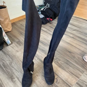 Svarta lårhöga stövlar med dragkedja - Säljer ett par svarta lårhöga stövlar med dragkedja på insidan. Skorna har en rund tå och är tillverkade i mocka. Perfekta för att lyfta vilken outfit som helst.