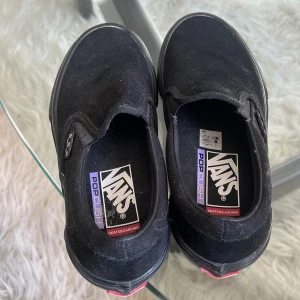 Svarta Vans PopCush slip-on skor - Svarta Vans slip-on med PopCush innersula, perfekt för skateboarding. Skorna har en klassisk låg profil, ovandel i mocka och den ikoniska gummisulan med rutmönster. Diskret Vans-logga på sidan och bekväm passform.