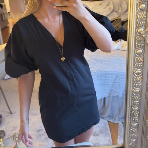 Svart kortklänning med puffärmar - Svart kortklänning med djup v-ringning och voluminösa puffärmar. Klänningen har en figurnära passform och är enkel men stilren, perfekt för dig som gillar minimalistisk design. Materialet känns mjukt och lätt.