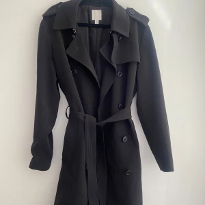Svart trenchcoat från H&M - Säljer en svart trenchcoat från H&M med klassisk krage, knappar framtill och bälte i midjan. Jackan har långa ärmar och en stilren design som passar perfekt till vår och höst. Enkel att matcha med olika outfits. Kappan har jag haft på mig en gång men råka fastna i något så den har fått en liten skavank. Den är inte alls tydlig men man kan säkert få bort den 🌸☺️