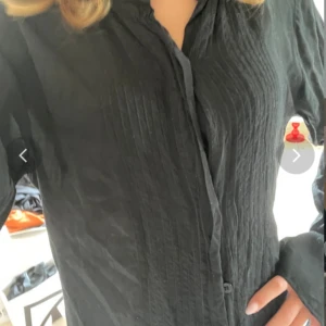 Svart blus med knappar och veckade detaljer - Säljer en svart blus med långa ärmar och knappar framtill. Blusen har fina veckade detaljer längs bröstet och en klassisk krage. Perfekt att styla till jeans eller kjol för en snygg look.