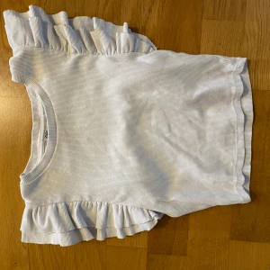 Vit ribbad topp med volang från Zara - Supersöt vit ribbad topp från Zara med volangdetaljer på axlarna. Perfekt för dig som vill ha något enkelt men ändå med en twist. Rund halsringning och korta ärmar med volang. Liten defekt (se bild) men syns knappast.