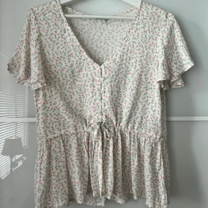 Blommig blus med knyt och volang - Söt blommig blus i vitt med små rosa och gröna blommor. Säljer då den tyvärr är för stor för mig. Bilderna är lånade men hör av dig om du vill ha egna!💕