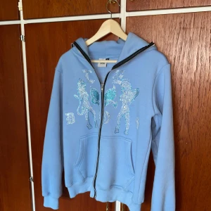 Ljusblå hoodie Rhinestone hoodie från Le Dos - Säljer en ljusblå hoodie från Le Dos med glittriga paljettdetaljer framtill och dragkedja. Tröjan har huva, fickor och långa ärmar. Perfekt för dig som vill sticka ut med en unik och snygg hoodie! Pris kan diskuteras vid snabb affär!