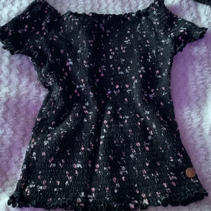 Svart smockad topp med blommigt mönster - Säljer en svart smockad topp med små rosa och vita blommor. Toppen har korta ärmar och volangkant nertill. Perfekt för dig som gillar stretchigt material och söt stil.