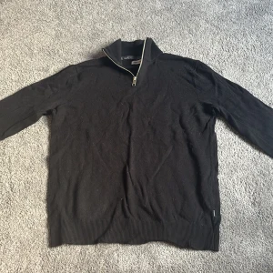 Svart tröja med dragkedja från Gant - Säljer en svart långärmad med halv dragkedja vid kragen. Tröjan har ribbade muddar vid ärmslut och nederkant, perfekt för en stilren och enkel look.