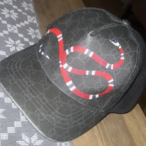 Svart keps från Gucci med ormtryck - Snygg svart keps från Gucci med diskret GG-mönster och en stor röd och vit orm på framsidan. Kepsen har mesh på baksidan för extra ventilation och justerbar passform. Perfekt för dig som vill sticka ut med en exklusiv accessoar.