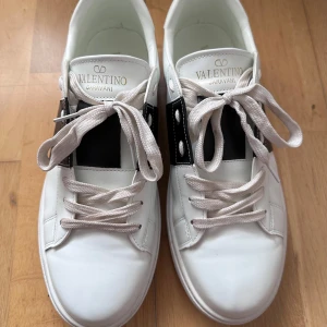 Vita sneakers Valentino Garavani - Snygga sneakers från Valentino Garavani med bred svart läderdetalj på sidan. Perfekta för dig som gillar stilrena och exklusiva sneakers. Använda 2 ggr men tyvärr lite för stora för min son. Köpta här på Plick. 