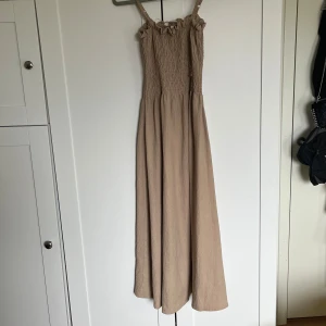Beige långklänning med smock och axelband - Säljer en beige långklänning med smock upptill och smala axelband. Klänningen har en luftig passform och är perfekt för varma dagar. Enkel och stilren design som passar till många tillfällen.