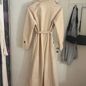 Beige trenchcoat från PrettyLittleThing - Snygg beige trenchcoat från PrettyLittleThing med klassisk krage, knappar och bälte i midjan. Lång modell med långa ärmar och axelklaffar. Perfekt för en stilren look. Aldrig använd och lappen finns kvar 