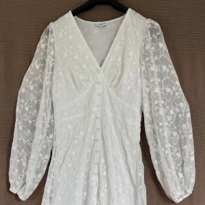 Vit blommig långklänning med v-ringning - Säljer en vit långklänning med v-ringning och långa puffärmar. Klänningen har broderade blommor, knappar framtill och är tillverkad i ett lätt, transparent material. Perfekt för sommarens speciella tillfällen.                    Använd 1 gång, djur- och rökfri. 