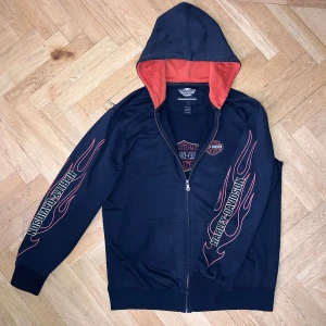 Svart hoodie från Harley-Davidson med flames - Säljer en svart hoodie från Harley-Davidson med dragkedja och orange insida på huvan. Ärmarna har tryckta flames och Harley-Davidson-logga. Tillverkad i 100% bomull och har fickor framtill.