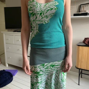 Grön och vit mönstrad långklänning - Säljer en långklänning med grönt och vitt blad- och randmönster. Klänningen har smala axelband och är ärmlös, med en figurnära passform upptill och en längre kjol. Perfekt för sommardagar!