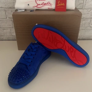 Blå sneakers från Christian Louboutin med nitar - Säljer ett par blå sneakers från Christian Louboutin med matchande blå nitar på tån och den ikoniska röda sulan. Skorna har snörning och är tillverkade i mocka med exklusiv känsla. Perfekta för dig som vill sticka ut med en lyxig och färgstark look.