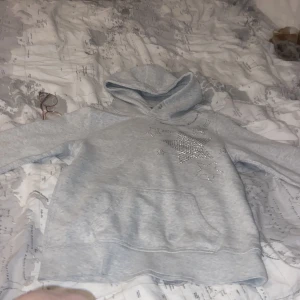 Ljusgrå hoodie med stjärnor från H&M - Säljer en ljusgrå hoodie från H&M med glittriga stjärnor framtill. Tröjan har huva och en stor magficka. Perfekt för dig som gillar en enkel men ändå lite extra look. Har en liten fläck på fickan men syns knappt.