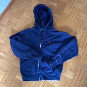 Marinblå hoodie från Polo Ralph Lauren - Säljer en marinblå hoodie från Polo Ralph Lauren med dragkedja och huva. Klassisk röd logga broderad på bröstet. Mjuk bomullskvalitet och två fickor framtill. Perfekt för en avslappnad stil.