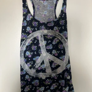 Blommig linne med peacemärke - Säljer ett svart linne med lila och blått blommönster och stort vitt peacemärke framtill. Linnet har brottarrygg och ärmlös design, perfekt för varma dagar.