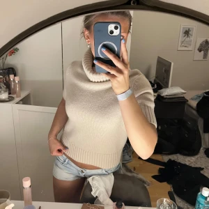 Beige stickad kortärmad polotröja - Säljer en beige stickad polotröja med korta ärmar. Tröjan har en hög, bred polokrage och är perfekt för lager-på-lager-stil. Enkel och stilren design som passar till mycket.