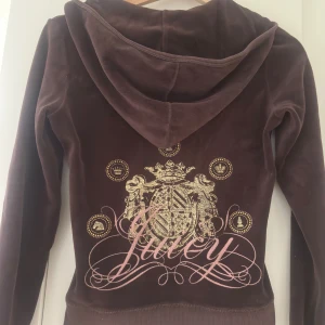 Brun velour hoodie från Juicy Couture - Mysig brun hoodie i velour från Juicy Couture med dragkedja framtill och huva. Baksidan har ett stort guldfärgat tryck med Juicy-logga, stl XS