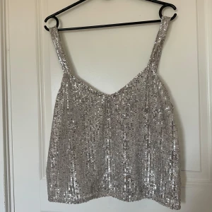 Silvrig paljettlinne - Säljer ett glittrigt silvrigt linne täckt av paljetter. Linnet har breda axelband och en rak, något croppad passform. Perfekt för dig som vill sticka ut och glänsa på festen!