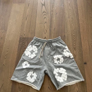 Grå shorts med vita blommor från Denim Tears - Gråa shorts från Denim Tears med vita blommiga tryck över hela plagget. Passar lite baggy. Pris kan diskuteras. Passar folk som har L i strlk oxå.