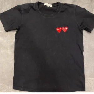 Svart t-shirt från Comme des Garçons - Säljer en svart t-shirt från Comme des Garçons med två röda hjärtan med ögon på bröstet. T-shirten har en klassisk rund hals. Storlek: M