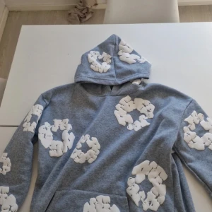 Grå hoodie denim tears - Säljer en grå hoodie med stora vita blommönster över hela tröjan och huvan. Tröjan har en klassisk huva och en magficka framtill. Perfekt för dig som vill sticka ut med en unik och bekväm stil. Denim tear