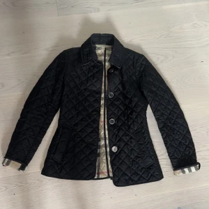 Burberry  - Säljer en klassisk svart quiltad jacka från Burberry i storlek XS. Jackan har knappar framtill, lång ärm och det ikoniska rutiga fodret. Snygg passform med markerad midja och dekorativ knappdetalj bak. Nu pris 9000-10000 säljer för 5000 pris kan ändrar vid bra affär😃