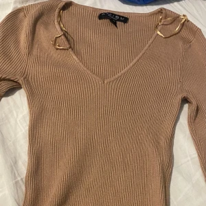 Beige ribbad tröja med v-ringning från Newyorker - Säljer en beige ribbad tröja från Newyorker med v-ringning och långa ärmar. Tröjan har dekorativa guldfärgade detaljer vid axlarna och är figurnära i modellen. Har använt ett par gånger 
