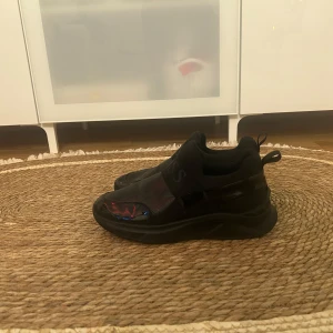 Svarta sneakers från BOSS med ormskinnsmönster - Säljer ett par svarta sneakers från BOSS med coolt ormskinnsmönster och bred resår över foten med BOSS-logga. Skorna har rund tå och platt sula, samt detaljer i blankt syntetmaterial. Perfekta för dig som vill ha något unikt och stilrent.