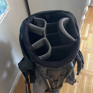 Svart och orange golfbag från DXT IZZO - Säljer en sportig golfbag från DXT IZZO i svart, grått och orange med flera fack och vadderade avdelare för klubbor. Väskan har bärrem och praktiska fickor för tillbehör. Perfekt för golfbanan och enkel att bära med sig.