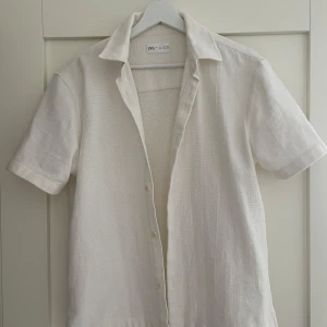 Vit kortärmad linneskjorta från Zara - Säljer en stilren vit skjorta från Zara i linne med korta ärmar och klassisk krage. Skjortan har knappar framtill och en luftig, lätt struktur som passar perfekt till varmare dagar. Enkel och tidlös design. Fantaskisk skick.