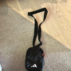 Svart axelväska från Adidas - Liten och smidig svart axelväska från Adidas med justerbart band. Väskan har dragkedja och en vit Adidas-logga på framsidan samt en röd detalj på dragkedjan. Perfekt för att bära med sig det viktigaste.
