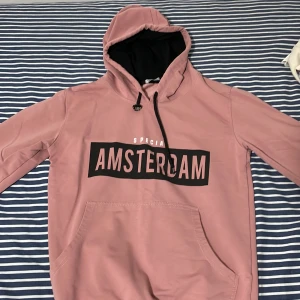 Rosa hoodie med tryck från NZR - Säljer en rosa hoodie från NZR med svart tryck där det står 'AMSTERDAM' på bröstet. Tröjan har huva med svart insida, dragsko och en stor magficka. Perfekt för en avslappnad stil.