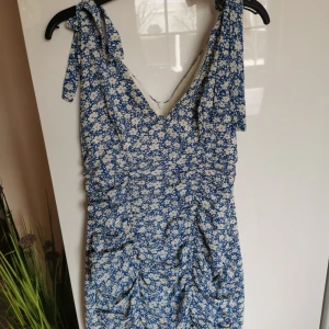 Blå blommig kortklänning med knytaxelband - Supersöt blå klänning med vita blommor och knytbara axelband. Djup v-ringning och rynkade detaljer framtill ger en snygg passform. Perfekt för varma dagar och somriga tillfällen.