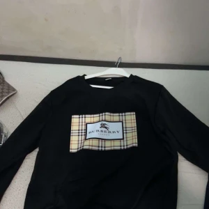 Svart långärmad tröja från Burberry - Svart långärmad tröja från Burberry med klassiskt rutigt mönster och logga på bröstet. Tröjan har rund halsringning och en stor beige, röd och svart rutig detalj framtill. Pris kan diskuteras vid snabb affär!