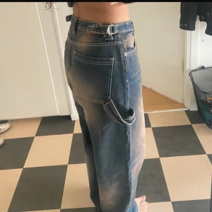 Baggy blå jeans - Säljer ett par baggy blå jeans med tvätt. Jeansen har låg/mellan midja, raka ben och tydliga kontrastsömmar. Endast använda 1 gång. Går att justera midjan med spännen på sidorna. 