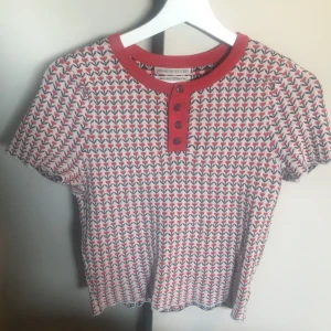 Mönstrad kortärmad topp från Urban Outfitters - Kortärmad topp från Urban Outfitters med röd och vit bas samt små mönster i hela tyget. Röd ribbad krage och knappslå med fyra röda knappar framtill. Perfekt för dig som gillar retrostil och färg.