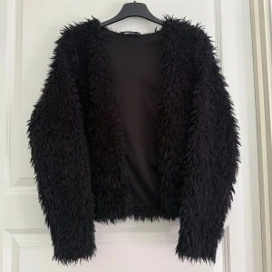 Svart pälsjacka från Gina Tricot - Säljer en svart fluffig pälsjacka från Gina Tricot. Jackan har lång ärm och är tillverkad i syntetmaterial med pälsliknande struktur. Perfekt för dig som vill sticka ut och hålla dig varm under kyliga dagar.