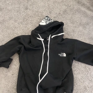 Svart hoodie från The North Face - Svart hoodie från The North Face med vit logga på bröstet och tryck i luvan. Tröjan har vit dragsko och känguruficka framtill. Perfekt för en avslappnad stil.