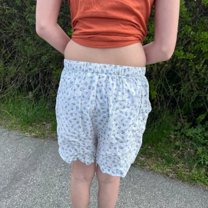 Blommiga shorts från H&M - Säljer ett par vita shorts från H&M med blått och grönt blommönster. De har elastisk midja och en lös passform, perfekta för varma dagar. Superbekväma och lätta att matcha med olika toppar.