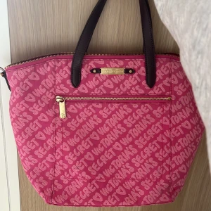 Rosa handväska från Victoria's Secret vintage - Säljer en rosa handväska från Victoria's Secret med mönster av loggan i ljusrosa. Väskan har smuts bruna handtag, guldfärgade dragkedjor och en praktisk ytterficka. Perfekt för att bära med sig allt man behöver.