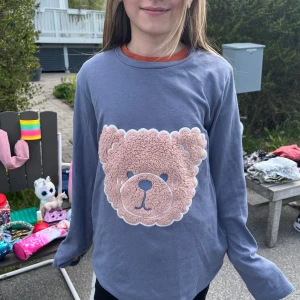 Ljuslila långärmad tröja med nalle från Tezenis - Säljer en ljuslila långärmad tröja från Tezenis med en stor, fluffig nalleapplikation framtill. Tröjan har rund halsringning med orange kant och är perfekt för dig som gillar gulliga detaljer.